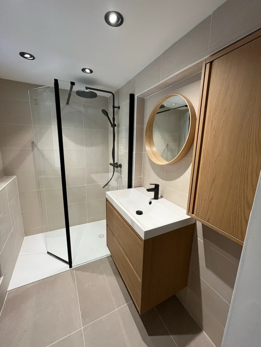 Modern bathroom with wood vanity, round mirror, white sink, black framed glass shower enclosure, and recessed ceiling lights